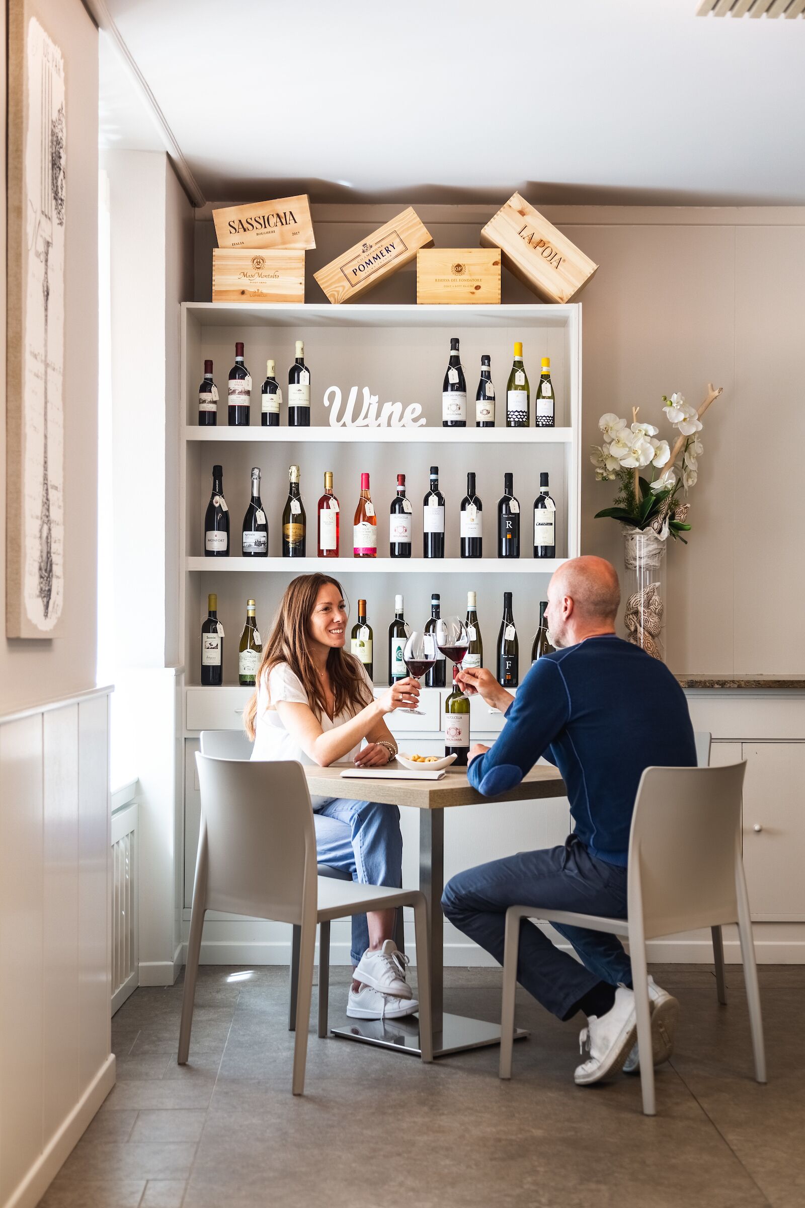 Un coin repas avec un meuble à vin où sont exposées différentes bouteilles, une table où sont assis deux personnes et un vase décoratif rempli de fleurs.