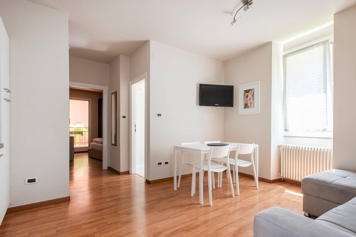Una sala de estar moderna con un sofá gris, una mesa de comedor blanca con sillas, un televisor de pantalla plana montado en la pared y un ventanal con persianas.
