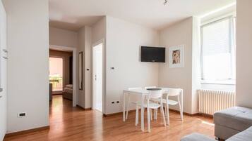 Una sala de estar moderna con un sofá gris, una mesa de comedor blanca con sillas, un televisor de pantalla plana montado en la pared y un ventanal con persianas.