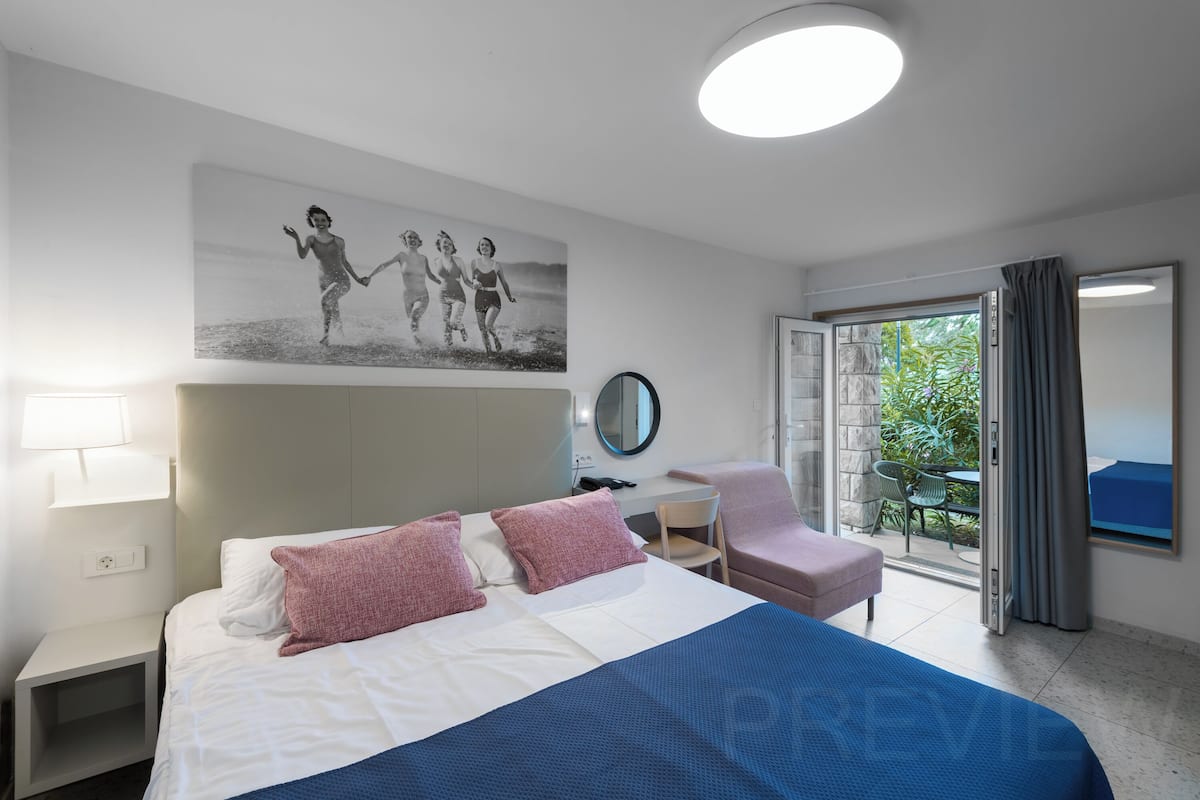 A hotel room with a bed, a desk, a chair, a mirror, and a balcony with curtains.
