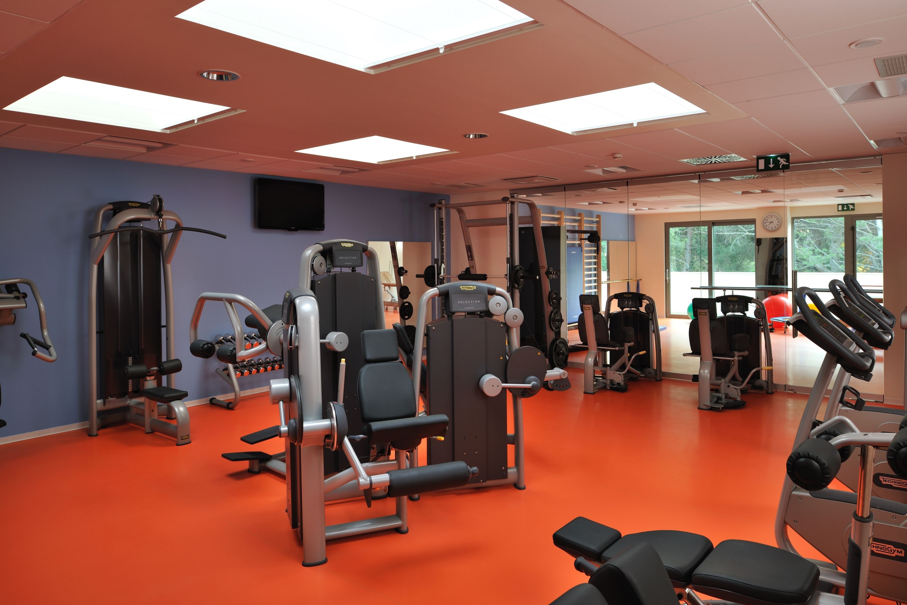 Une salle de sport équipée de divers appareils d’exercice, avec une horloge et une fenêtre donnant sur des arbres.
