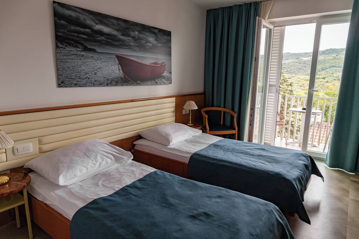 Une chambre d’hôtel avec deux lits, une table de chevet, une lampe, une chaise, un balcon avec vue et un tableau représentant un bateau accroché au mur.