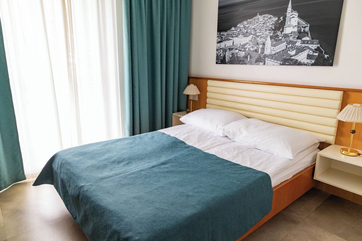 Un lit bien fait, recouvert d’une couvre-lit turquoise, avec un oreiller blanc et une tête de lit en bois. Une table de chevet avec une lampe et un tableau représentant un paysage urbain sont posés à côté du lit.