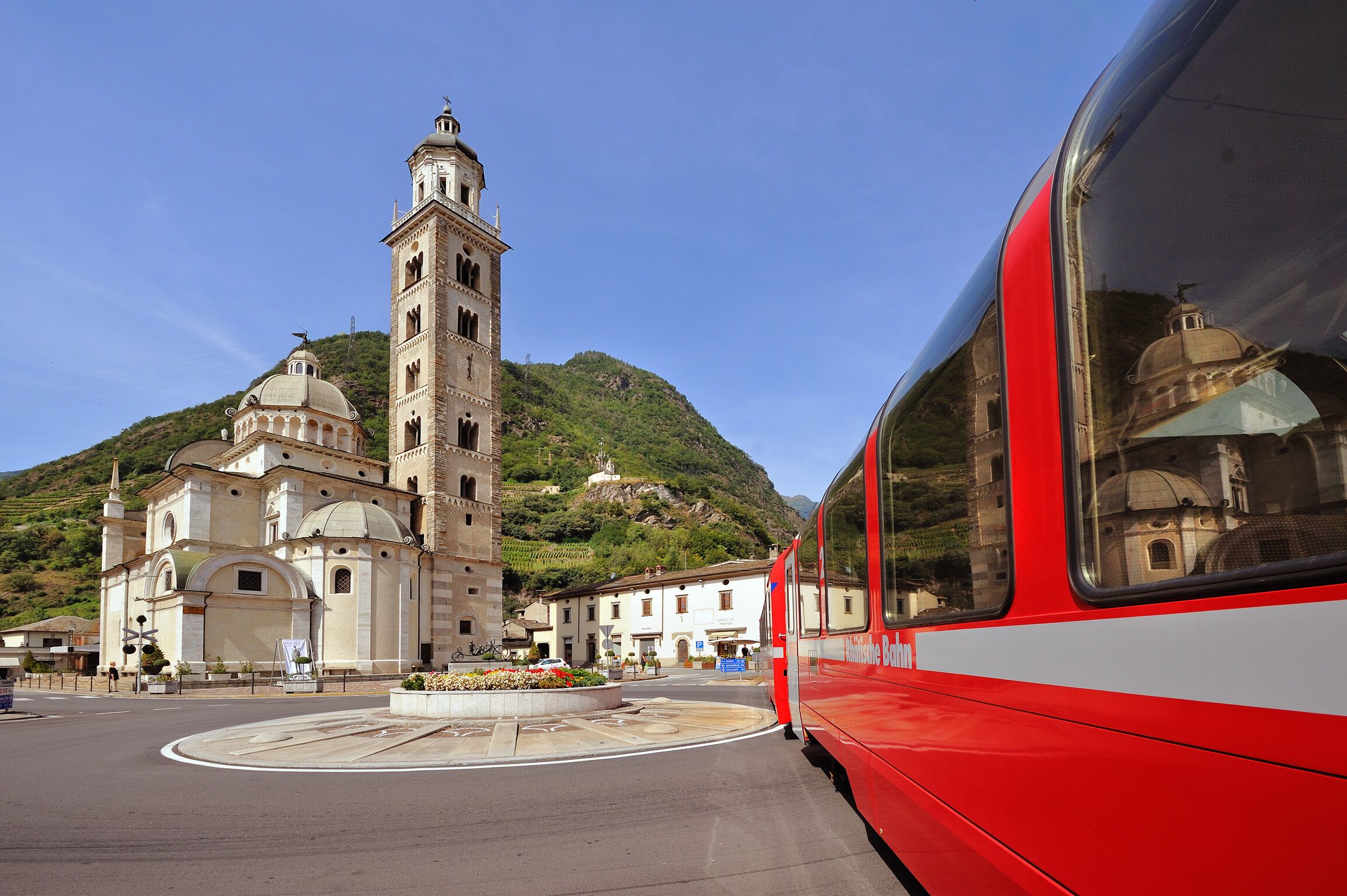 A red bus parked in front of a historic church with a bell tower and dome, surrounded by a hilly landscape.