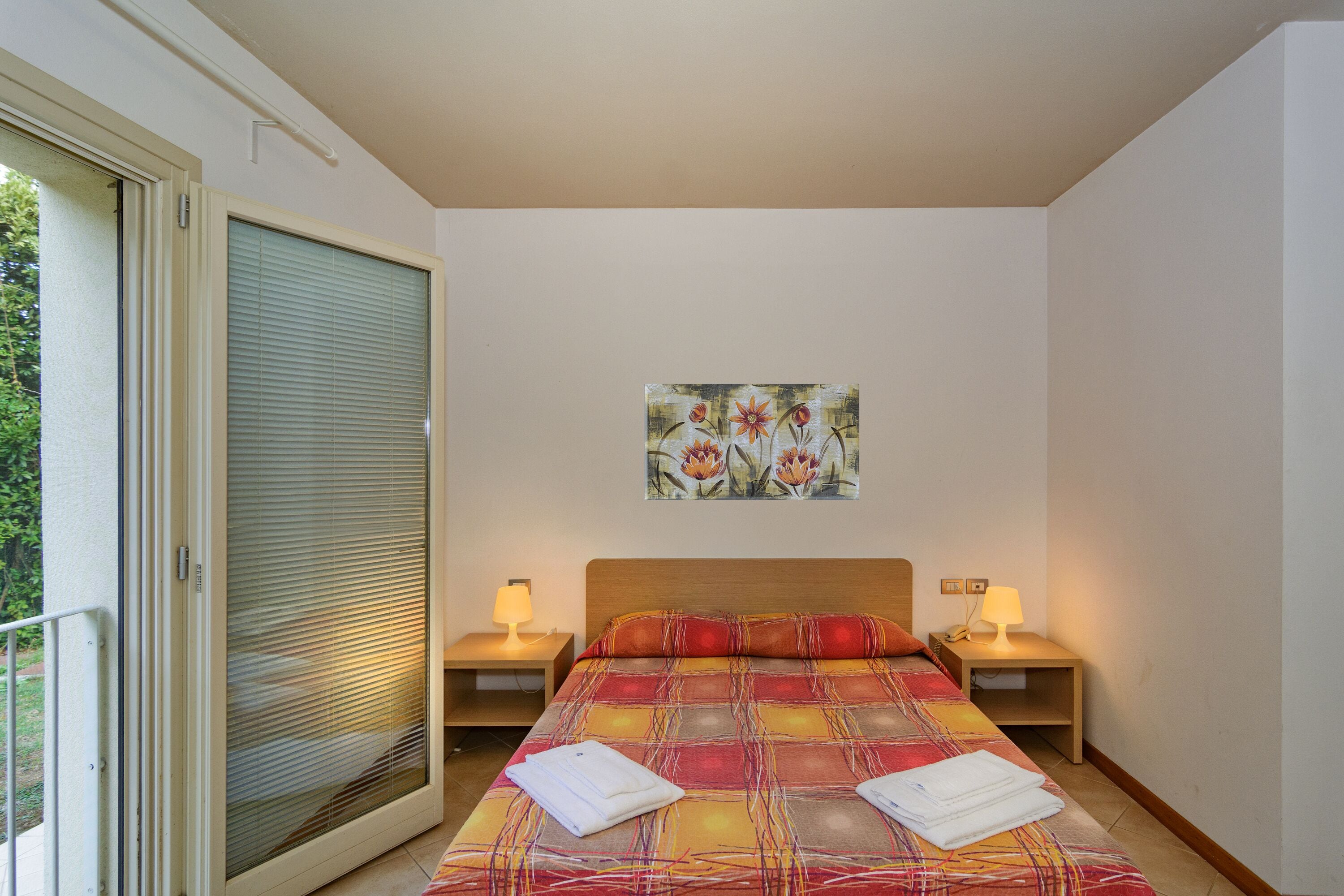 A bedroom with a large bed, two bedside tables with lamps, a window with blinds, and a painting on the wall.