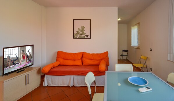 Una sala de estar con sofá, televisor mostrando una clase de yoga, una mesa de comedor con sillas y un ventanal pequeño.