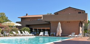 A pool area with lounge chairs and umbrellas, a brick building with a flat roof, and a clear blue sky.