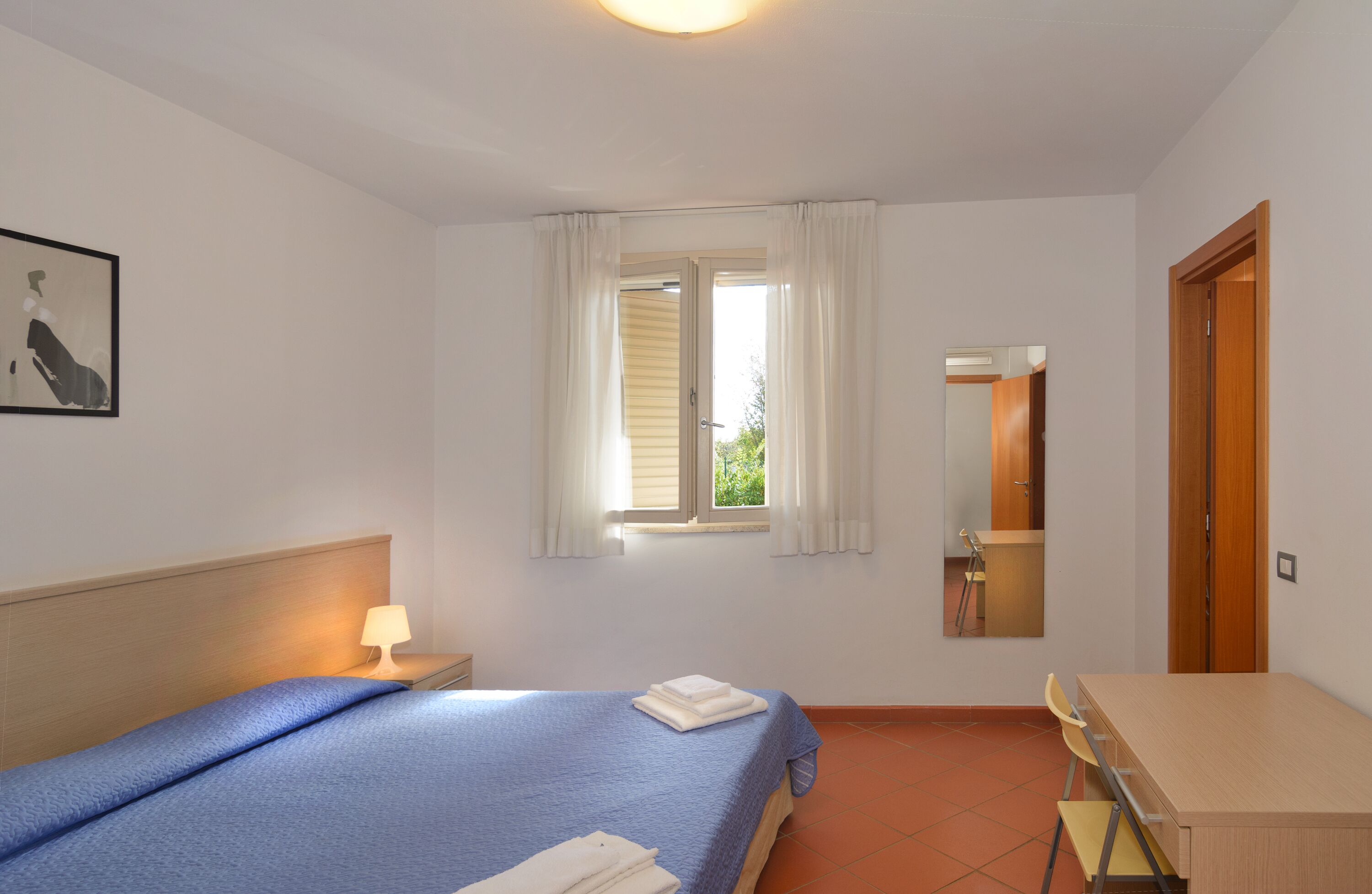 Une chambre à coucher comprenant un lit, un bureau, une chaise, une fenêtre avec des rideaux, une lampe de chevet et un miroir.