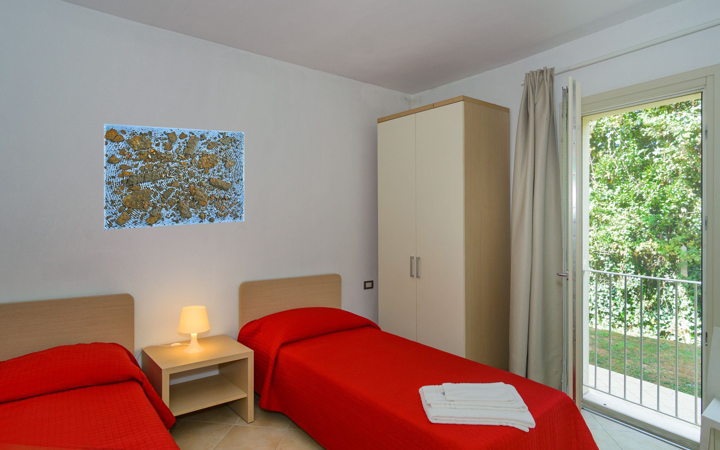 Een kamer met twee bedden, een kledingkast, een nachtkastje met een lamp en een raam met uitzicht op groen.
