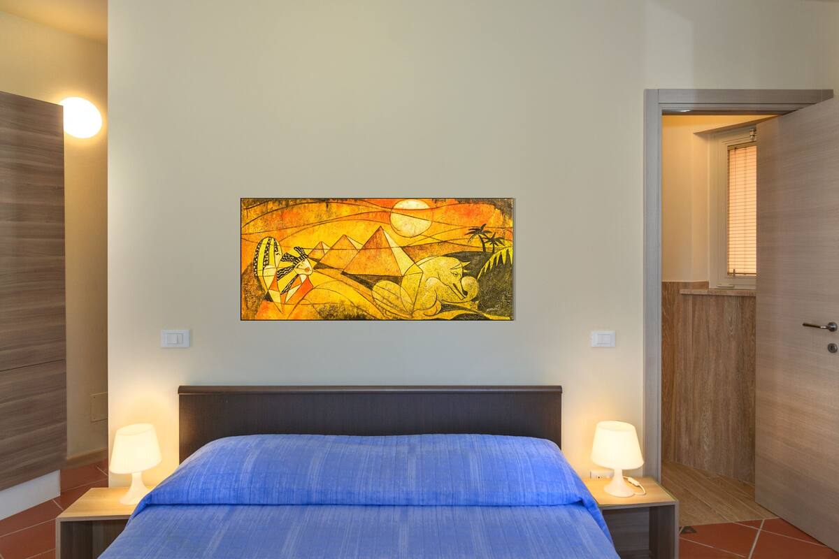 A bedroom with a bed, two bedside tables with lamps, a wall-mounted painting, and a door leading to another room.