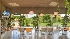 A modern indoor dining area with a view of a garden. There are tables and chairs arranged for dining, and potted plants are placed around the room.