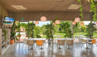 A modern indoor dining area with a view of a garden. There are tables and chairs arranged for dining, and potted plants are placed around the room.