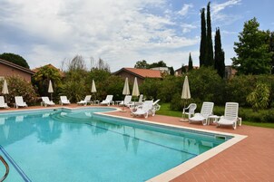 A swimming pool with white lounge chairs and umbrellas, surrounded by greenery and buildings.