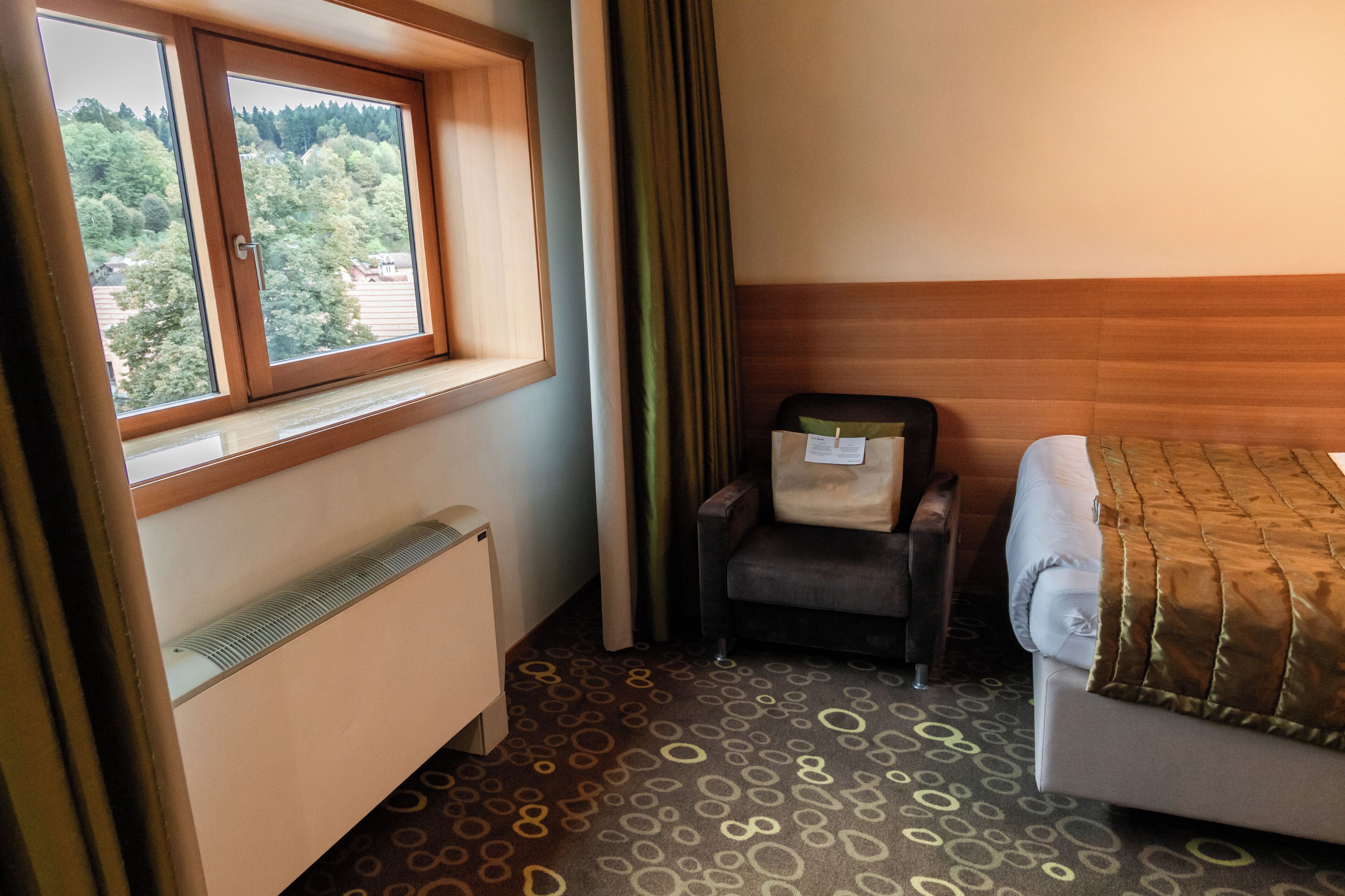 A hotel room with a bed, a chair, a radiator, a window with a view of trees, and a patterned carpet.