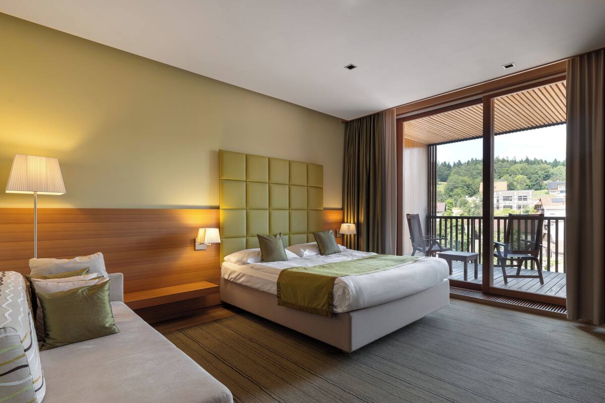 A hotel room with a large bed, a sofa, a balcony with a view of the city, and a wooden headboard.