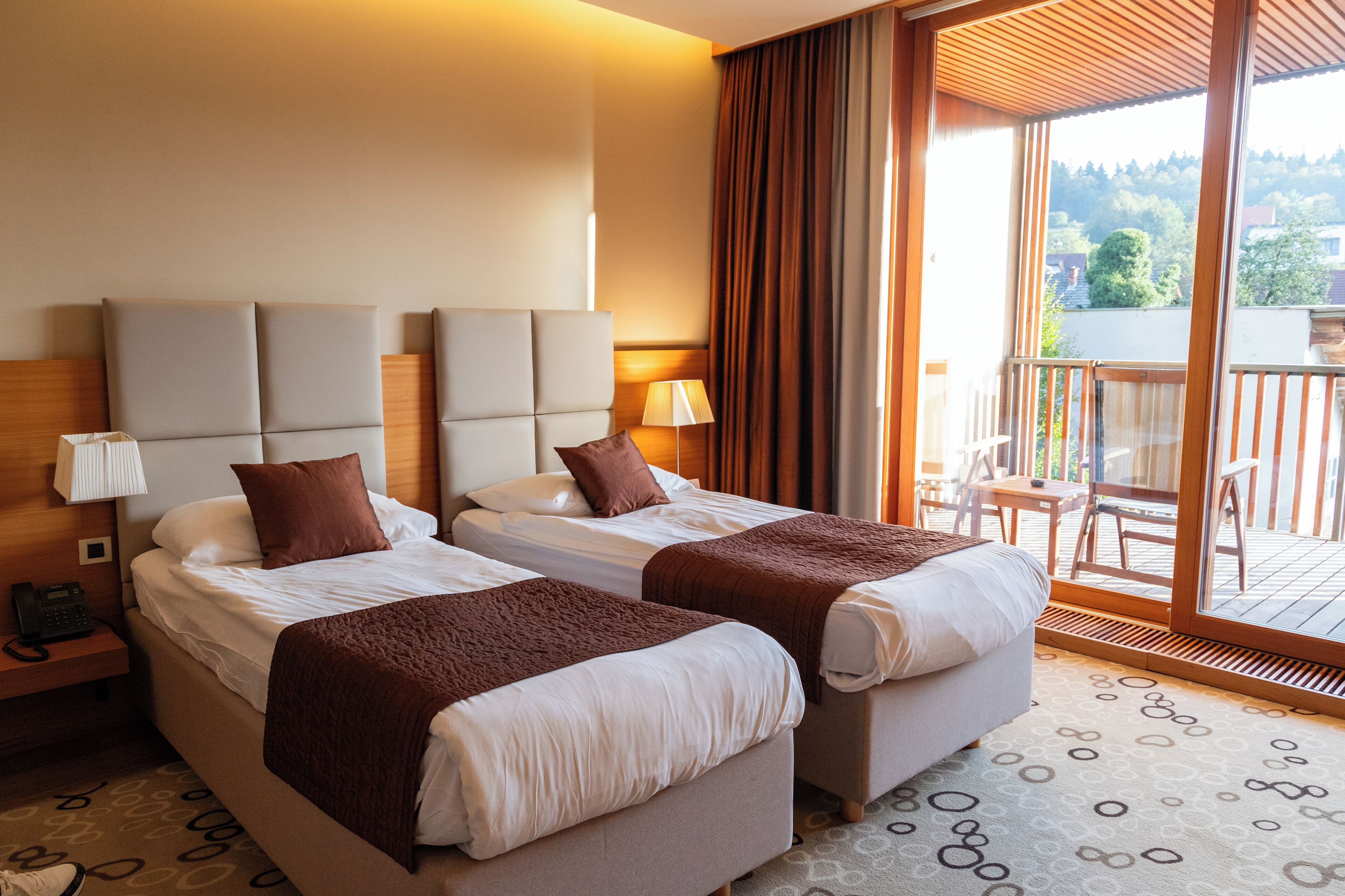 A hotel room with two beds, a nightstand with a phone, a lamp, and a view of the balcony.