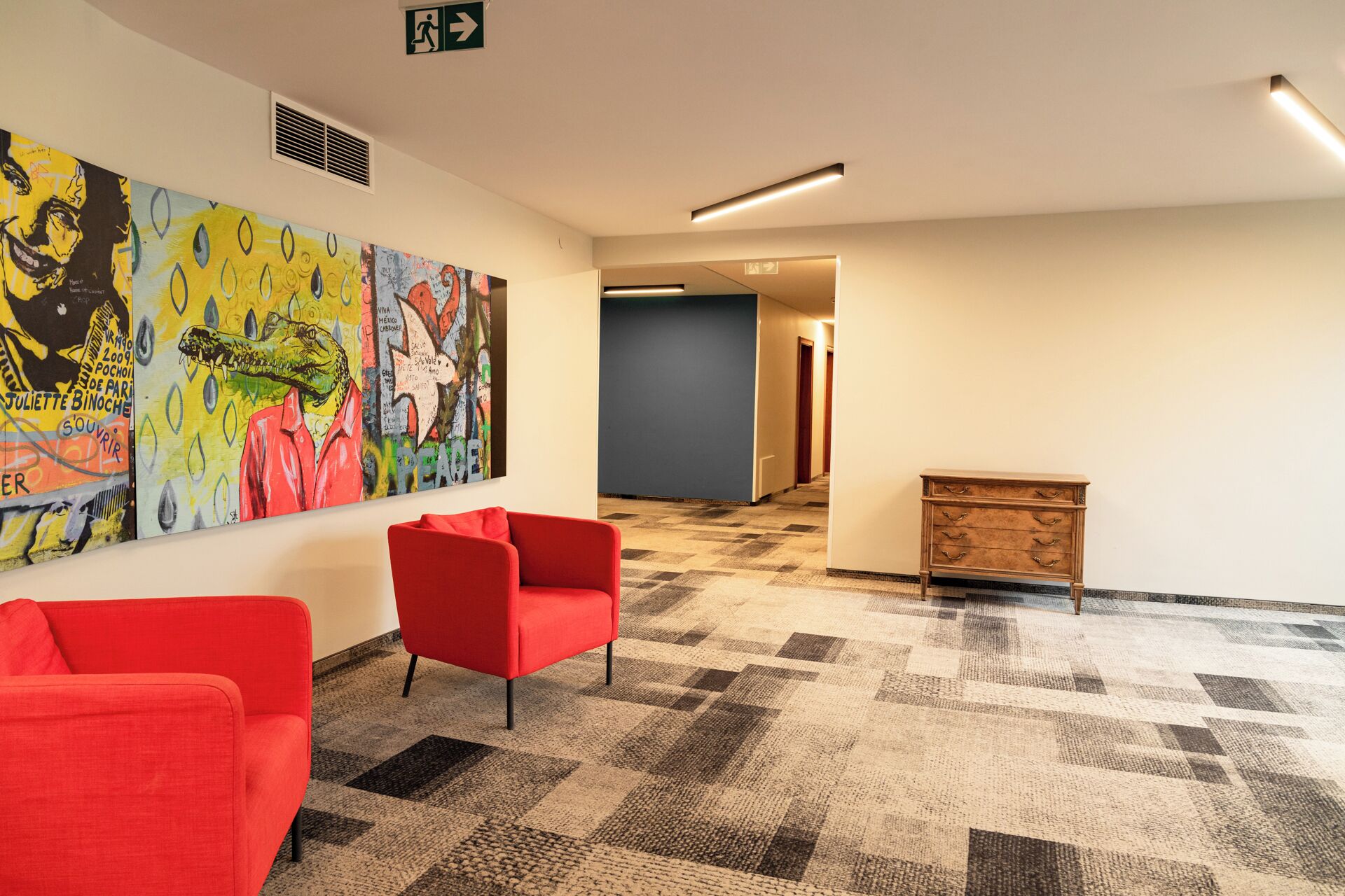 Een moderne lobby met een geruit tapijt, rode stoelen, een houten kast en kleurrijke abstracte kunst aan de muur.