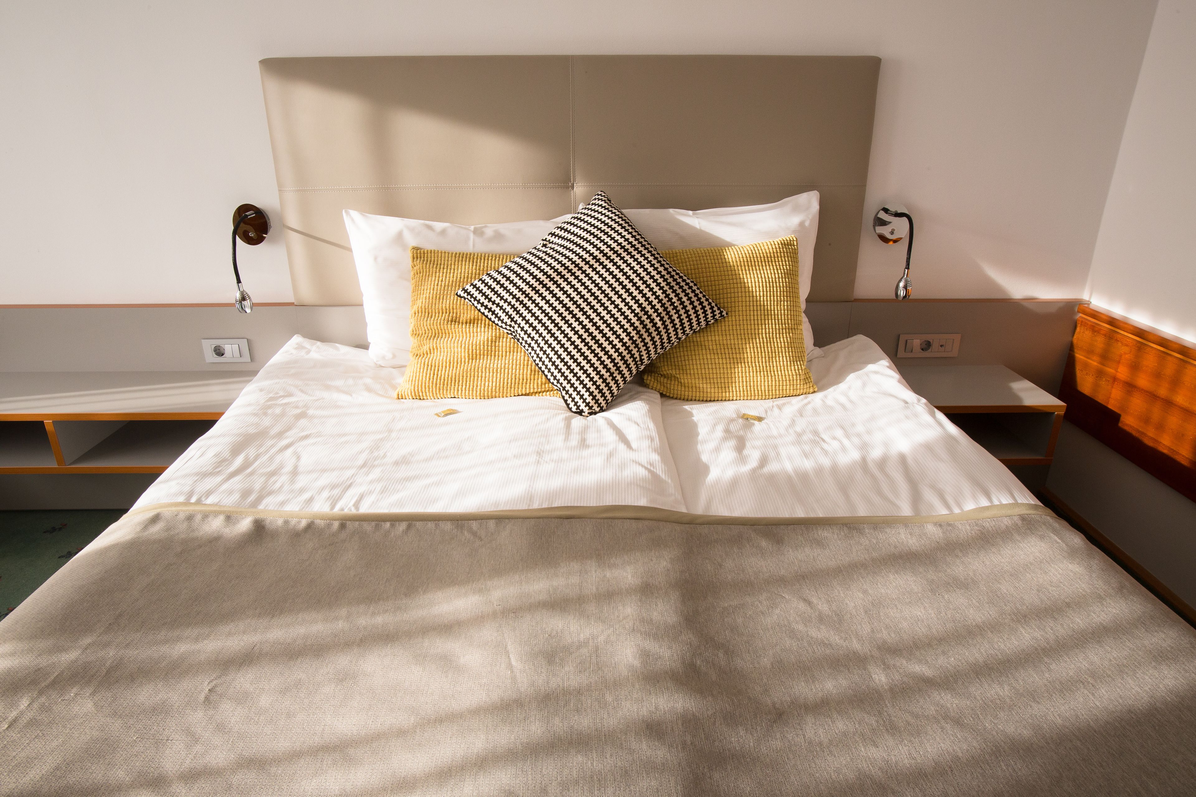 Una cama bien tendida con un cabecero color beige, dos almohadas y una mesita de noche con una lámpara.