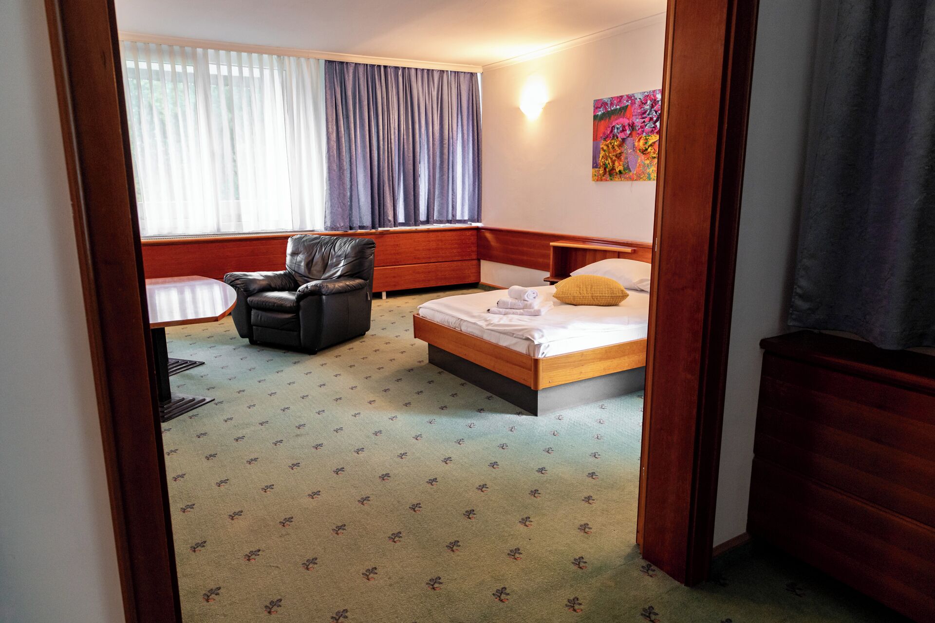 A hotel room with a bed, a chair, a small table, and a window with curtains.