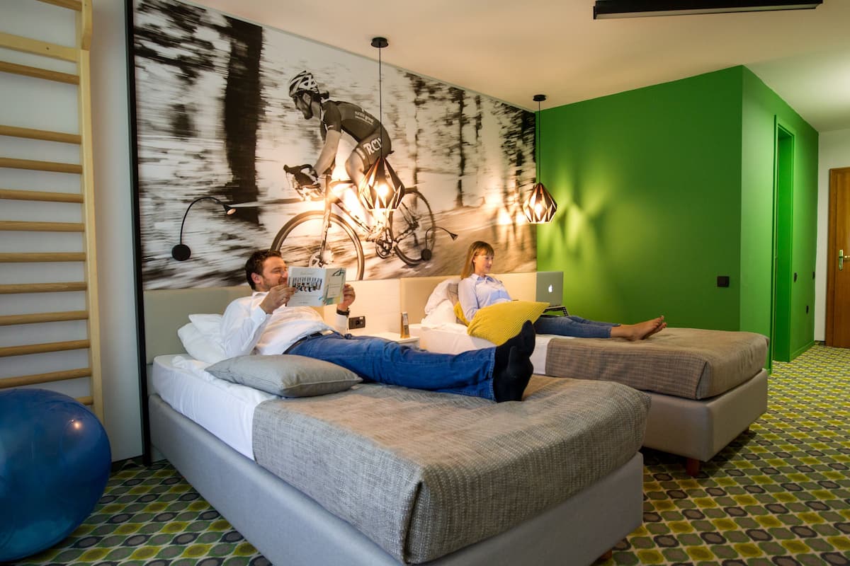 A room with two beds, a large wall mural of a cyclist, a green accent wall, and patterned flooring.