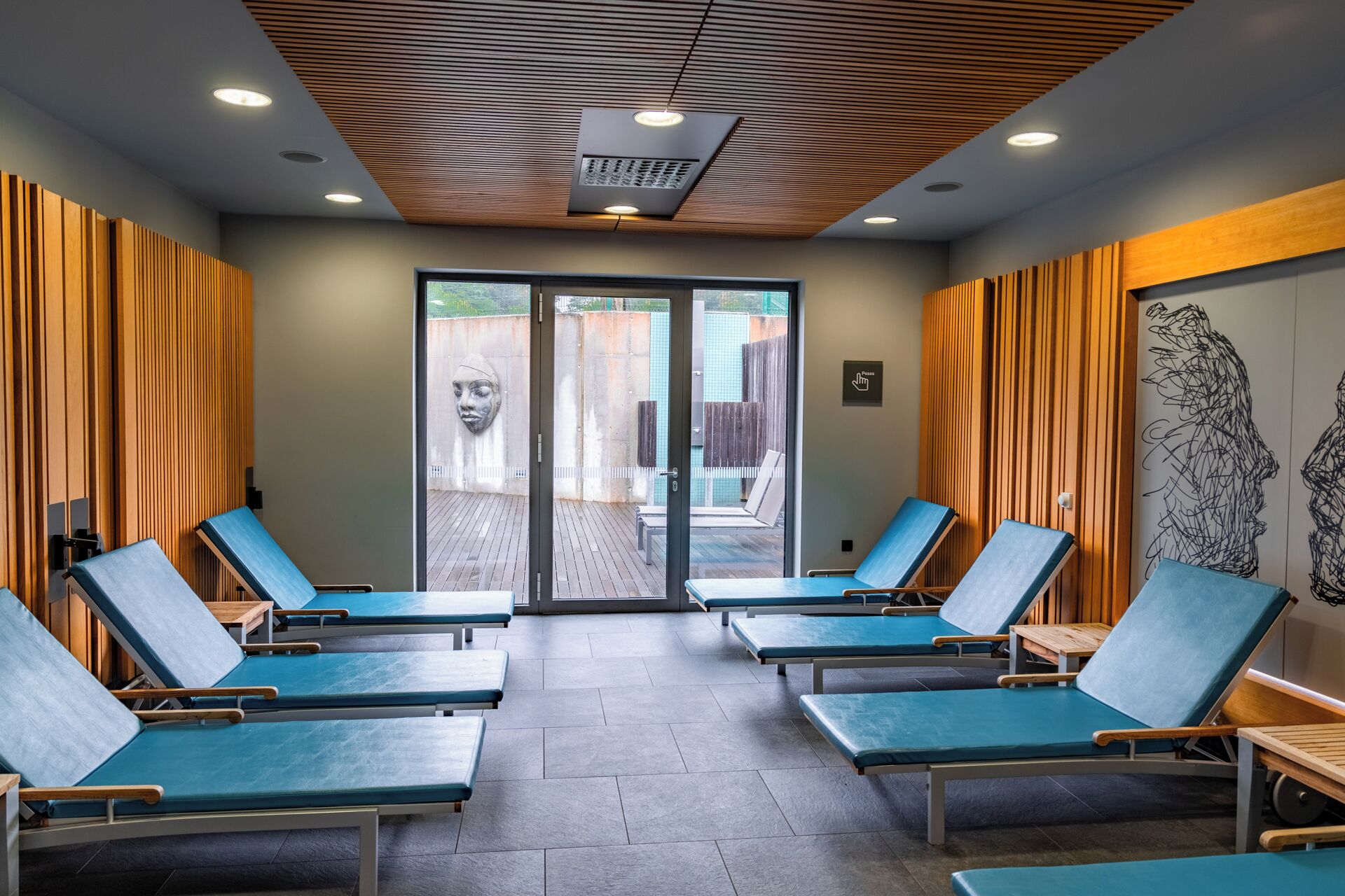 A spa room with four blue massage tables, wooden panel walls, and a large glass door leading outside.