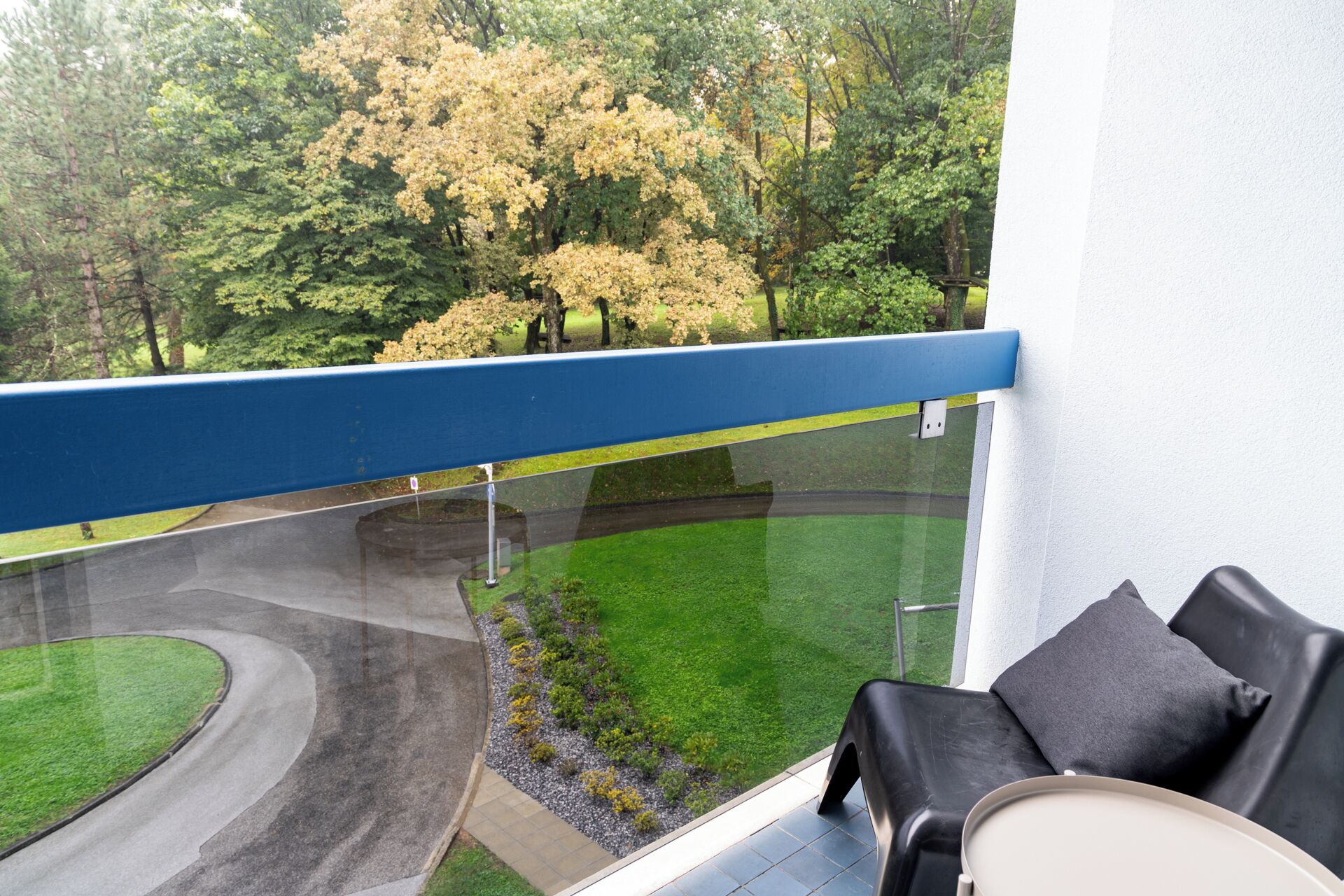 A balcony with a curved glass railing overlooking a landscaped area with trees and a winding path.