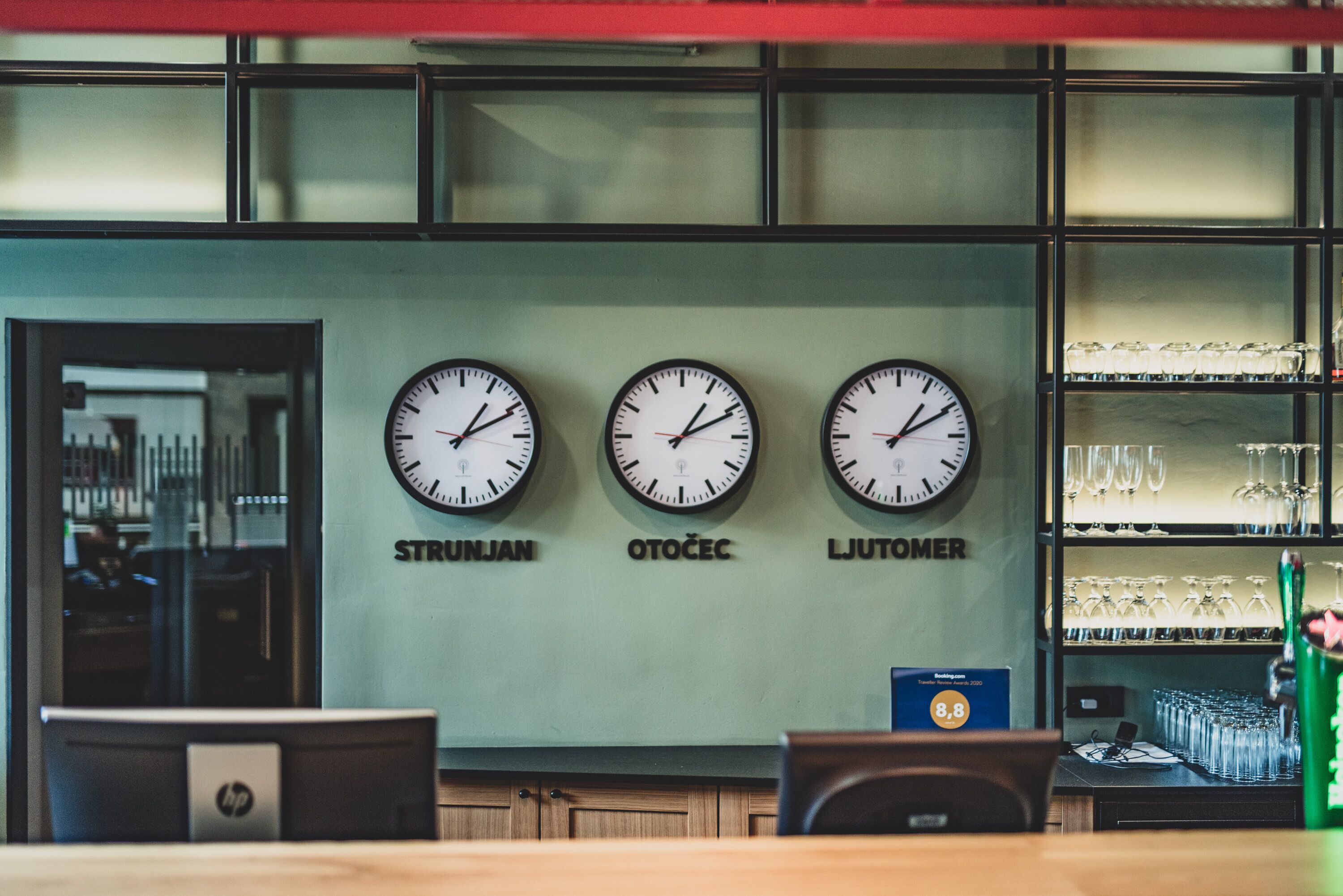 A reception area with three clocks labeled "STRUNJAN," "OTOČEC," and "LIUTOMER," and a glass display case with various items.