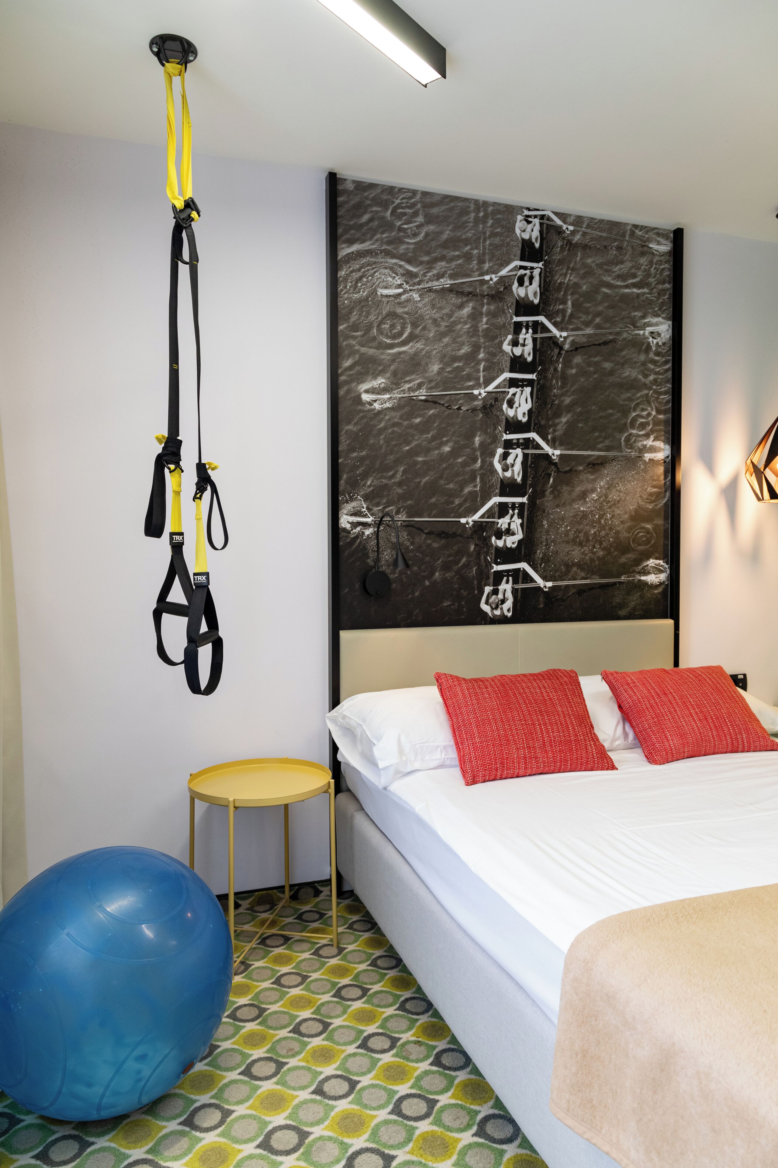 Une chambre avec un lit, un appareil d’entraînement de suspension TRX, un ballon d’exercice bleu et une œuvre d’art accrochée au mur.