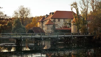 Un bâtiment historique avec un toit rouge, une tour en pierre et un pont enjambant une étendue d’eau calme.