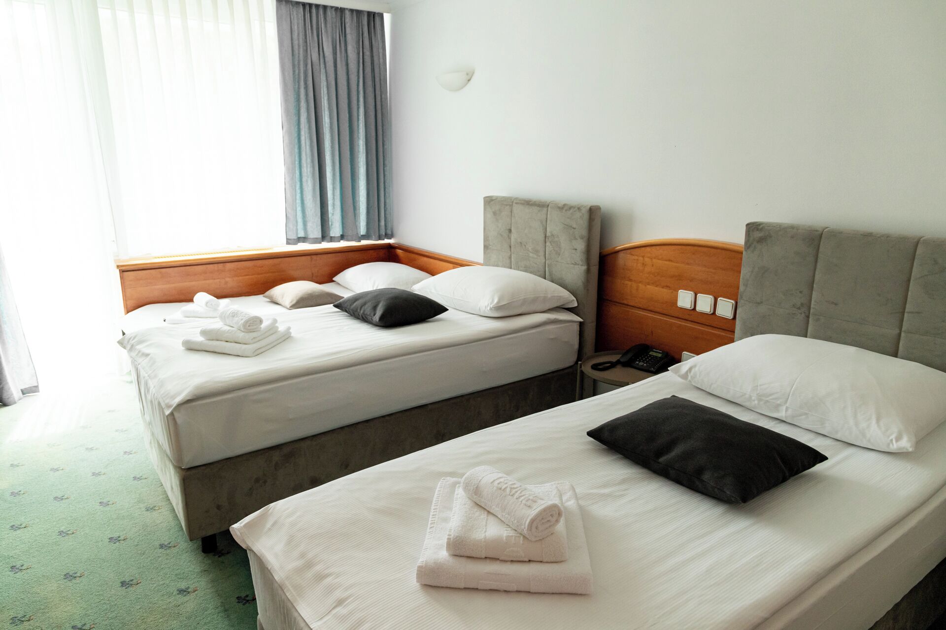 Habitación de hotel con dos camas, cada una con ropa blanca y almohadas, un cabecero de madera, una mesita de noche con teléfono y una ventana con cortinas.