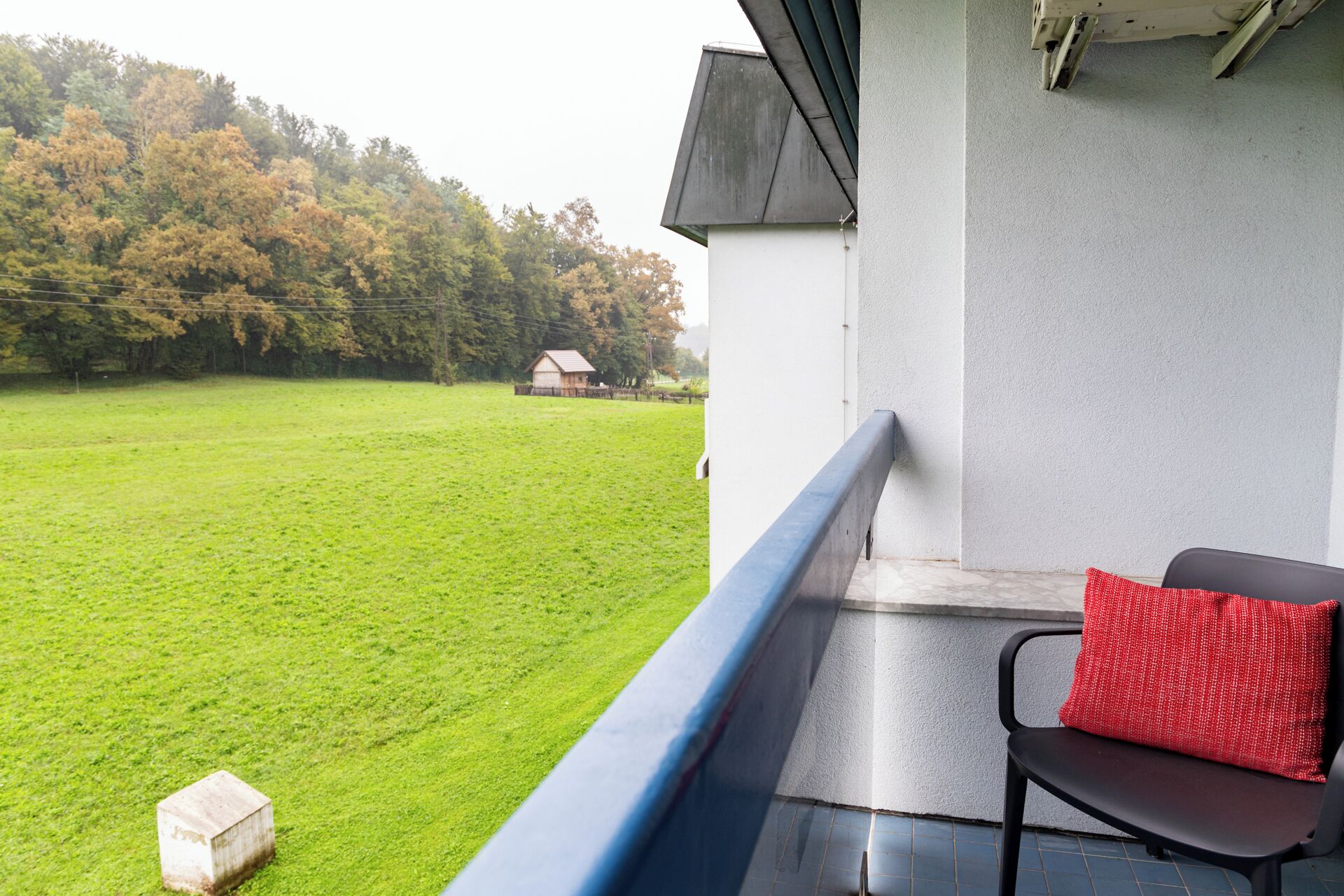 A balcony with a view of a green field and trees, a small house in the distance, and a red cushion on a chair.