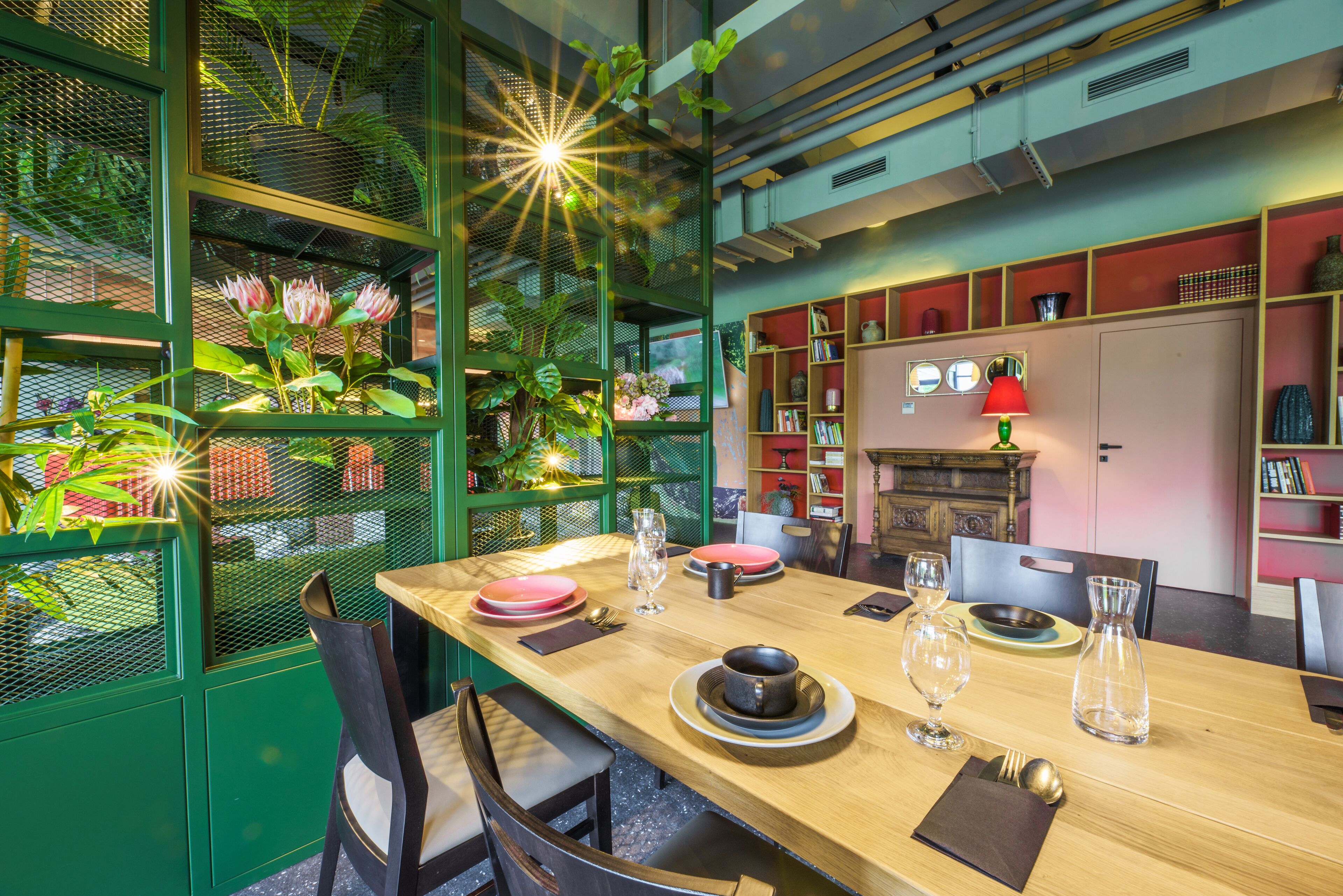 A dining area with a wooden table set for four, surrounded by chairs. There are plants in a green mesh enclosure to the left, and a red lamp on the table.
