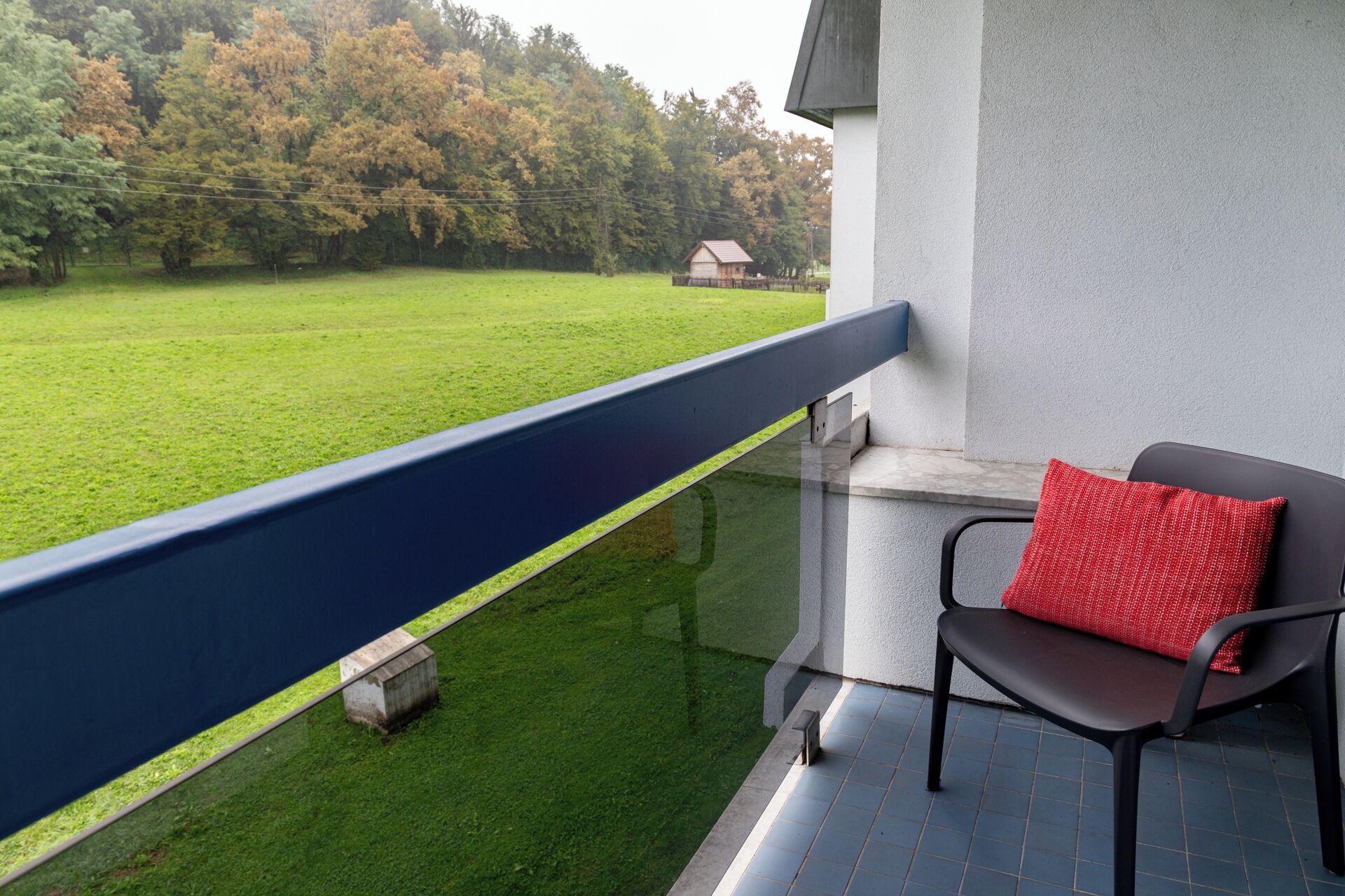 A balcony with a view of a green lawn and trees, a small house in the distance, and a chair with a red cushion.