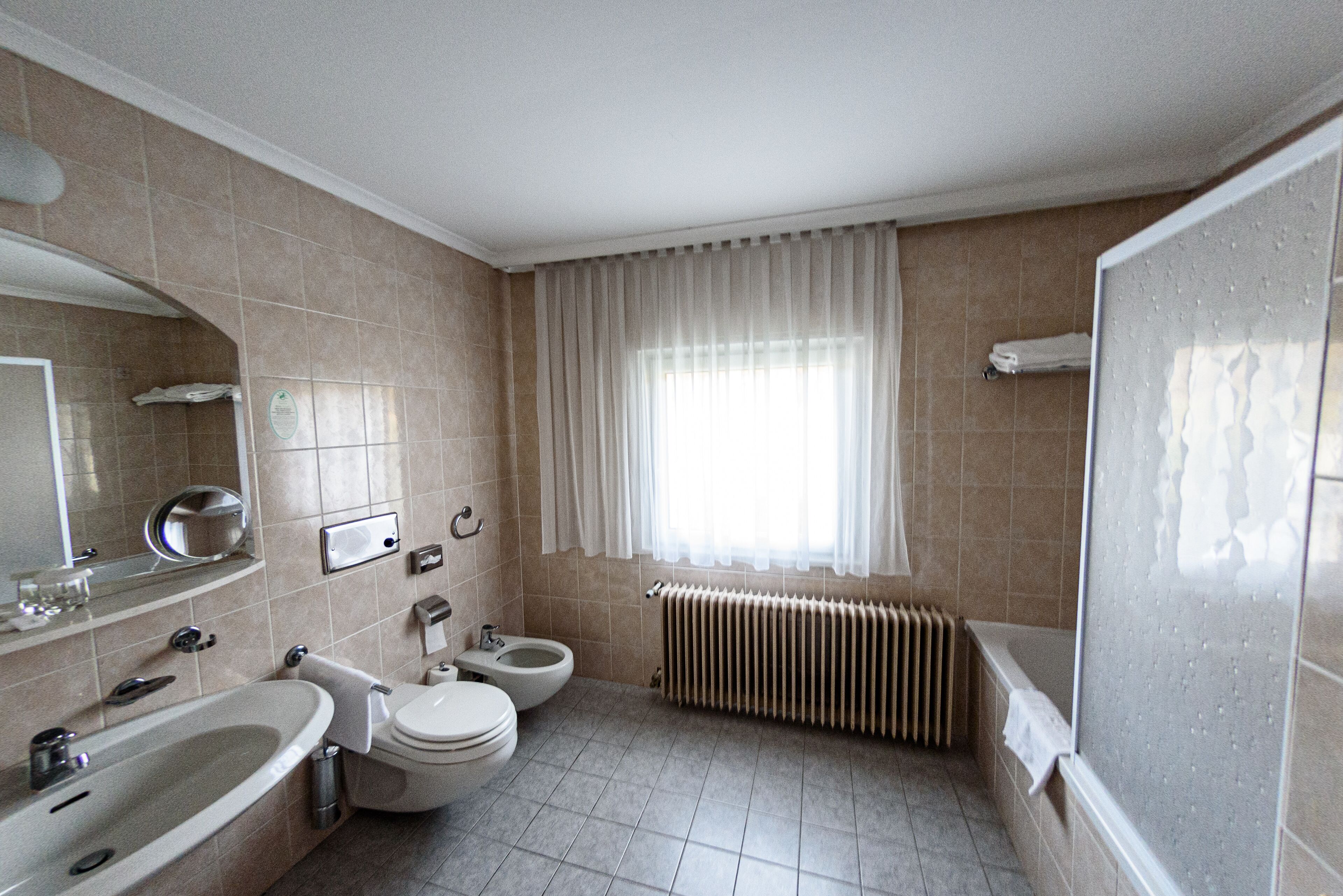 Et badeværelse med vask, toilet og bidét, fliserede vægge, et vindue med gardiner og en bruseplads.