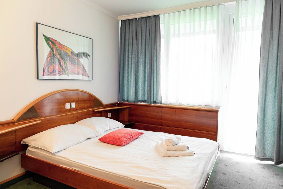 A hotel room with a wooden bed, white bedding, a red pillow, a framed artwork on the wall, and a window with curtains.