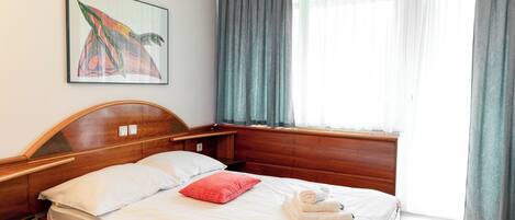 Habitación de hotel con cama de madera, ropa de cama blanca, almohada roja, una obra de arte enmarcada en la pared y una ventana con cortinas.