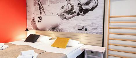 A hotel room with a bed, a small table, a wall-mounted shelf, and a large poster of a sailing scene.