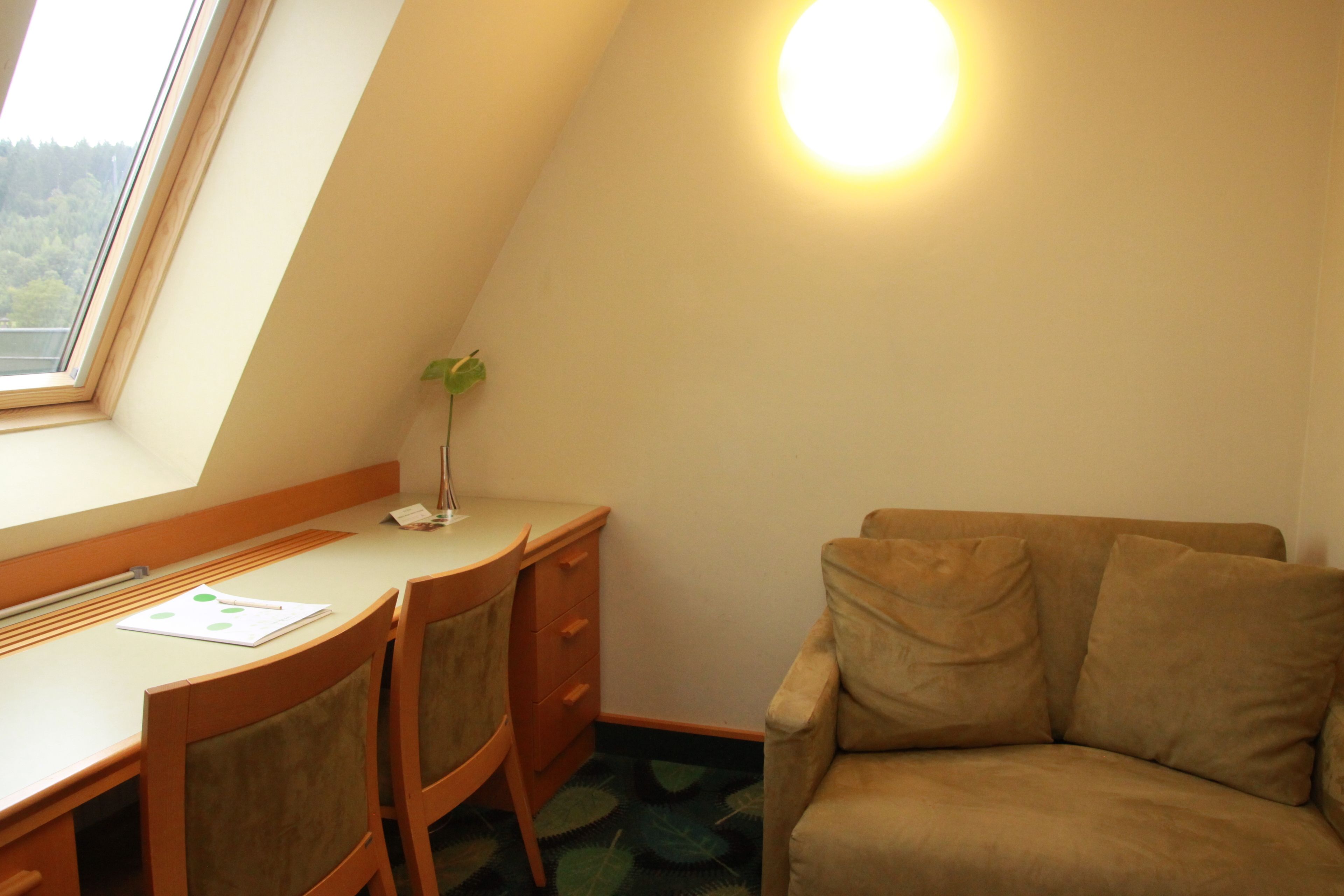 A room with a desk, two chairs, a small plant, a sofa, and a window with a view of trees.