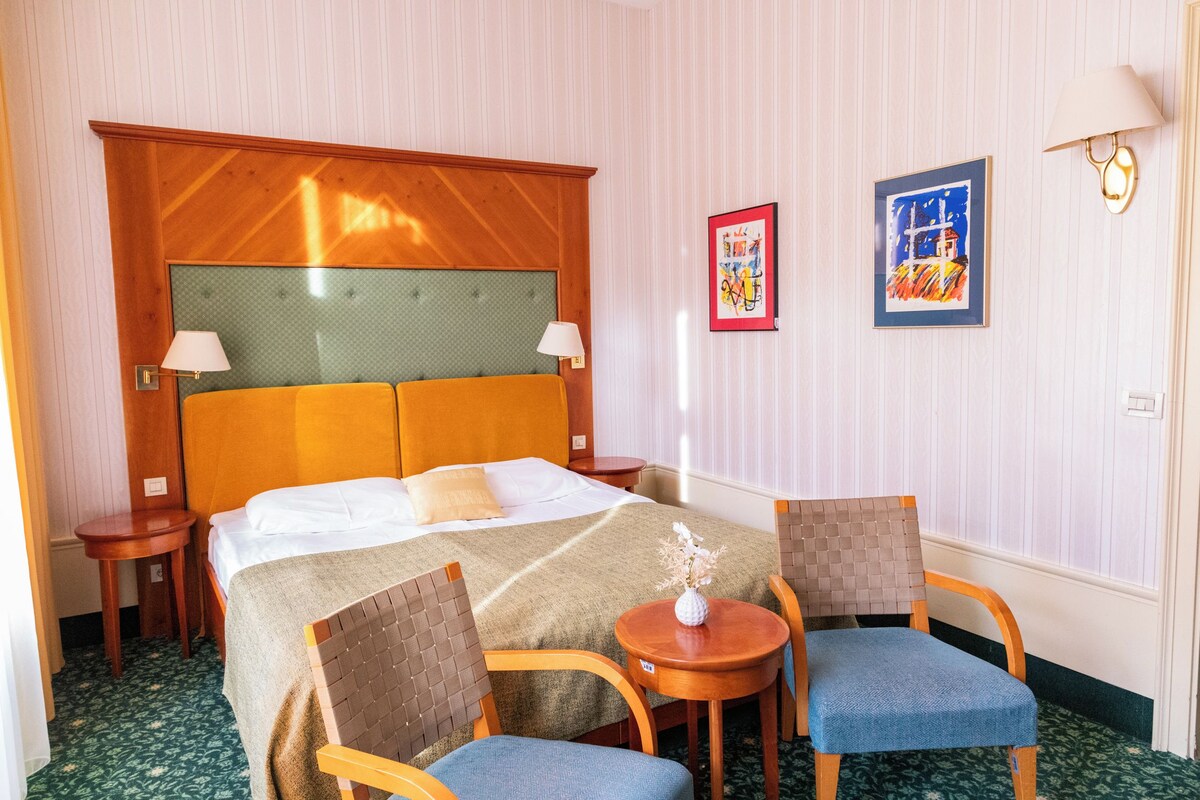 A hotel room with a bed, two chairs, a small table, a lamp, and two framed pictures on the wall.