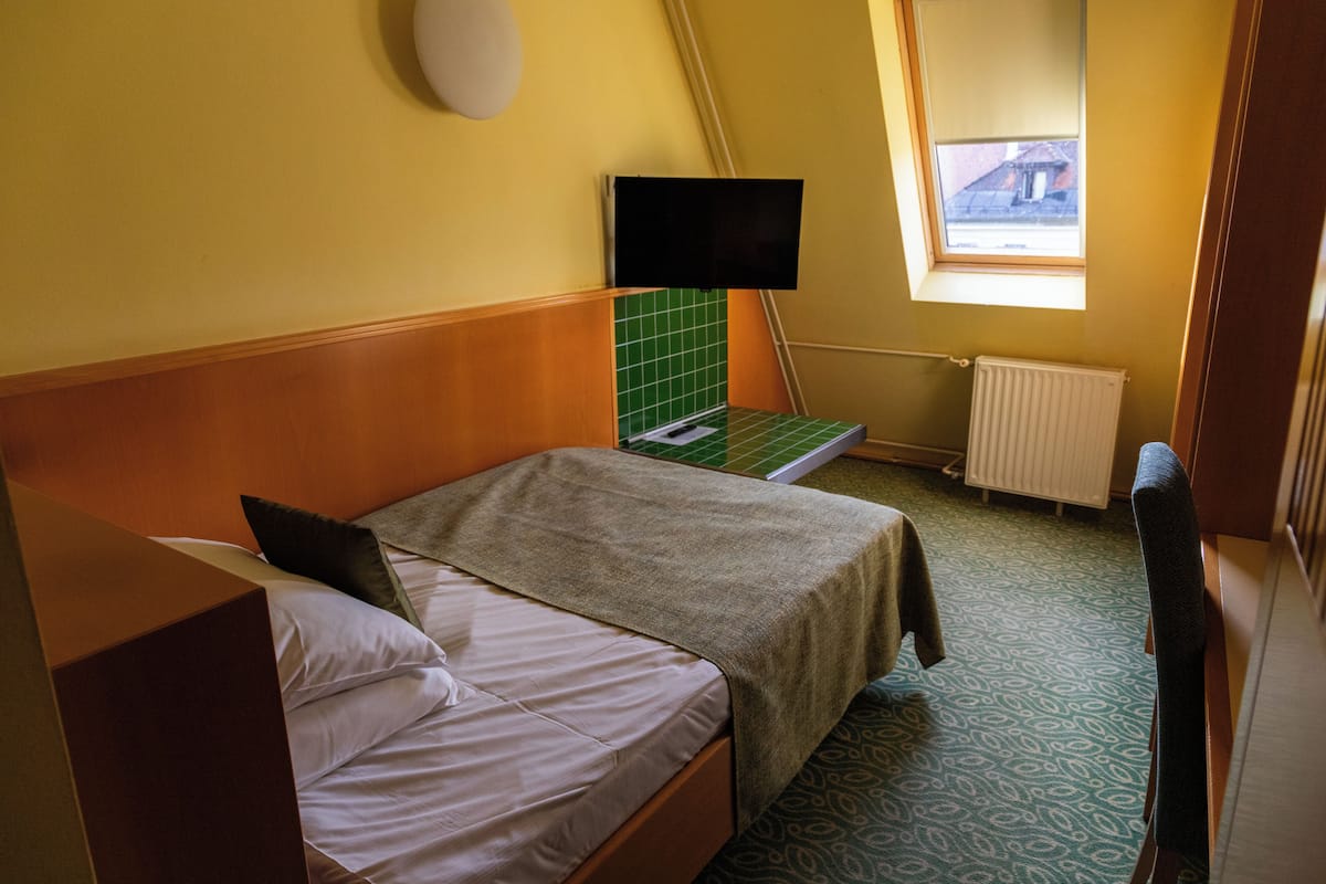 A small, single-bed room with a wooden headboard, a green tiled accent wall, a television mounted on the wall, and a window providing a view of the outside.