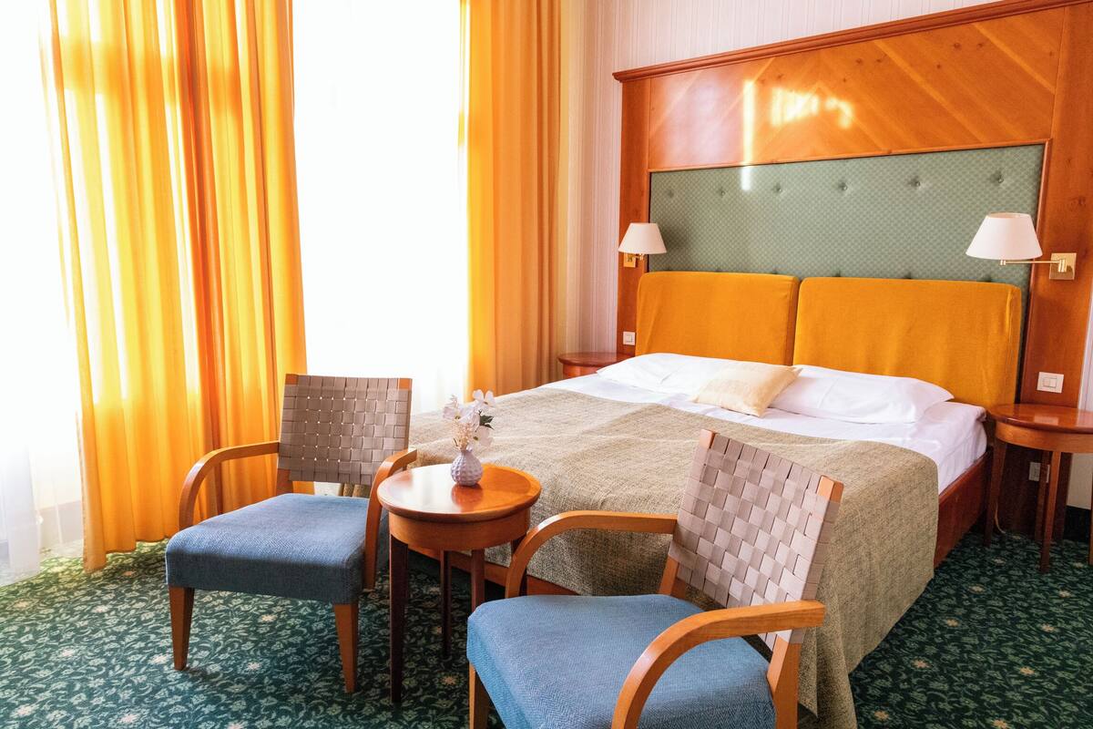 A hotel room with a large bed, two bedside tables with lamps, a small table with a vase, two chairs, and a patterned carpet.