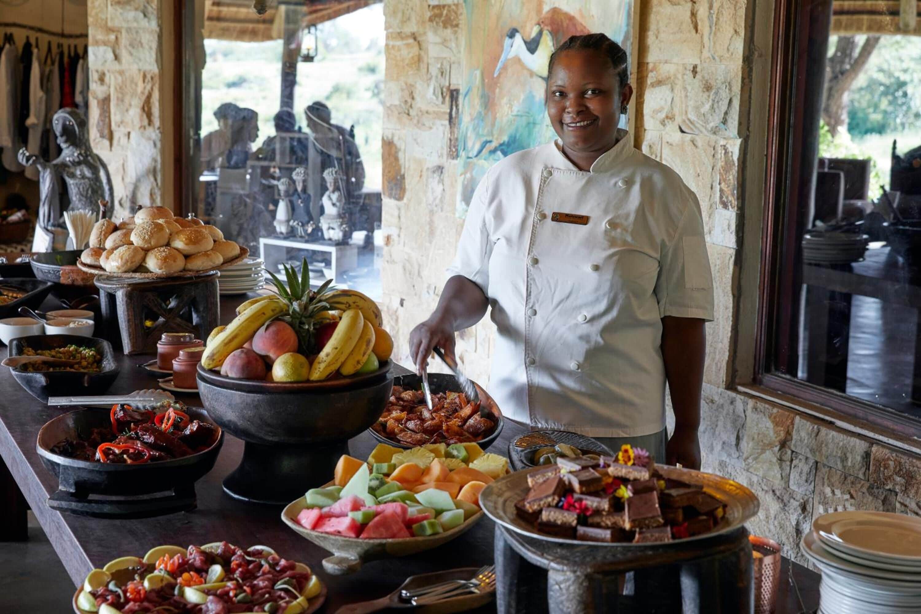 Un chef con uniforme blanco de pie detrás de una mesa de buffet con diversos platos, entre ellos frutas, carnes y postres.