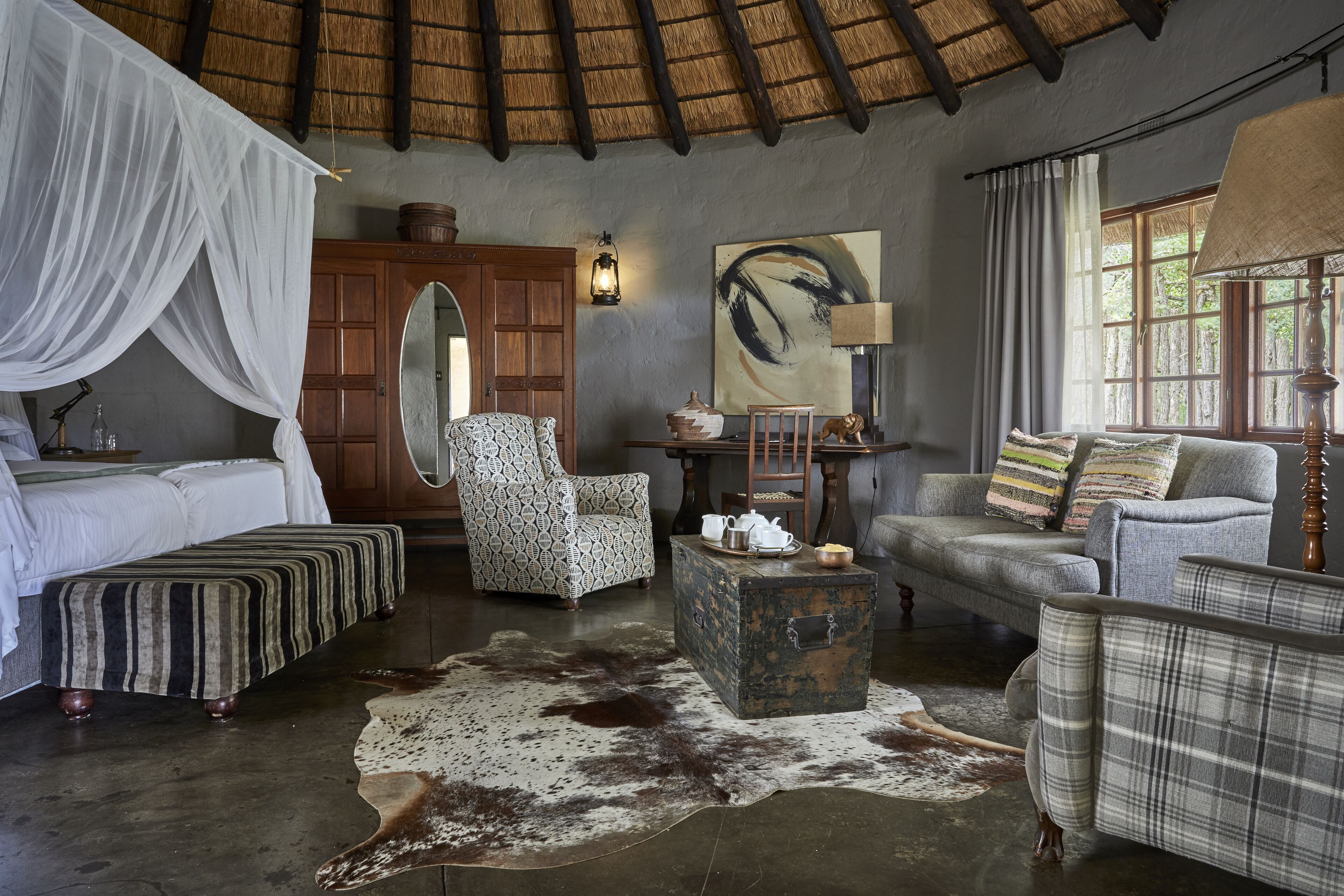 A room with a bed, a patterned armchair, a sofa, a wooden chest, and a cowhide rug.
