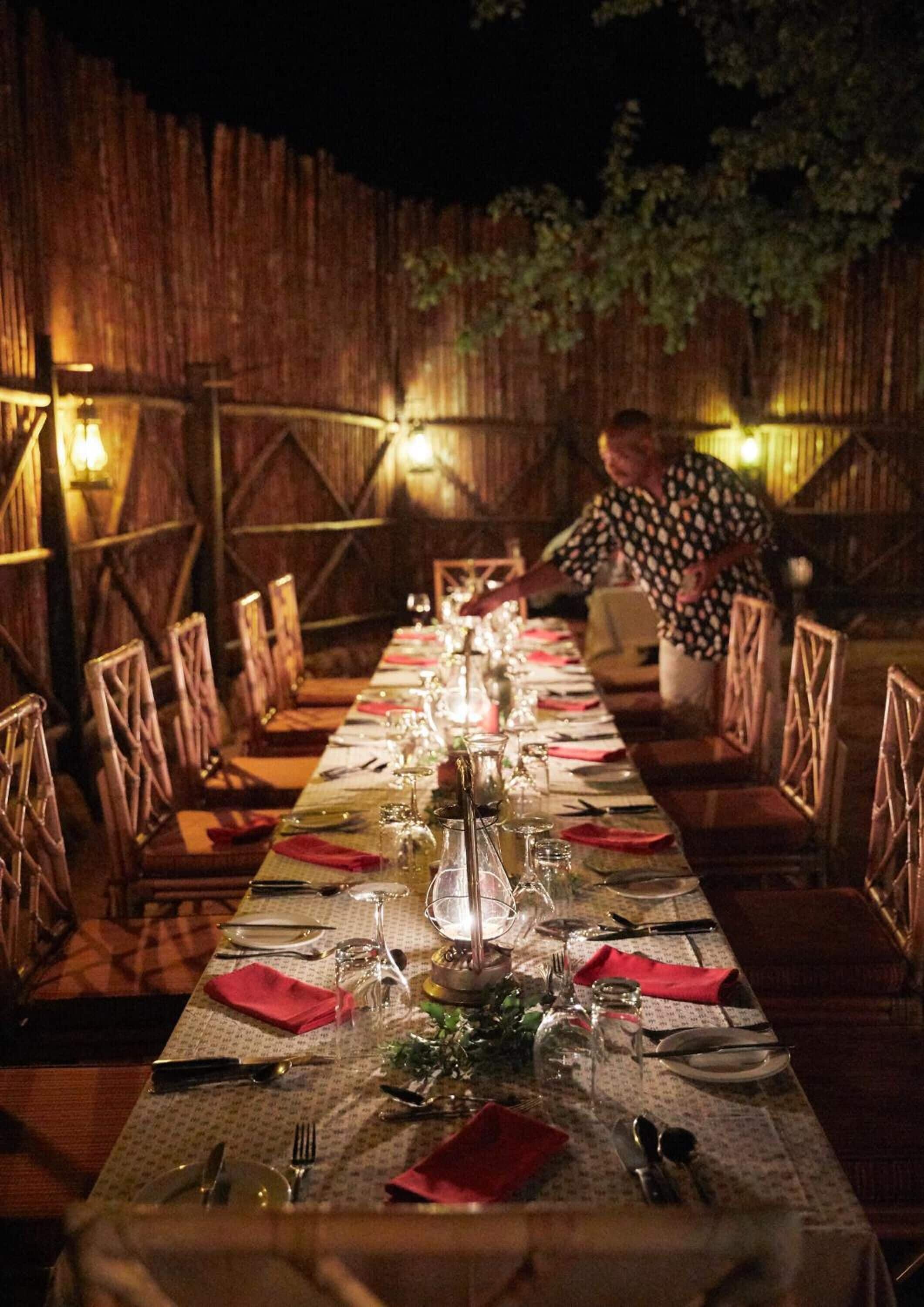 A long dining table set with red napkins, glassware, and plates, surrounded by wooden chairs in an outdoor setting with a wooden fence and warm lighting.