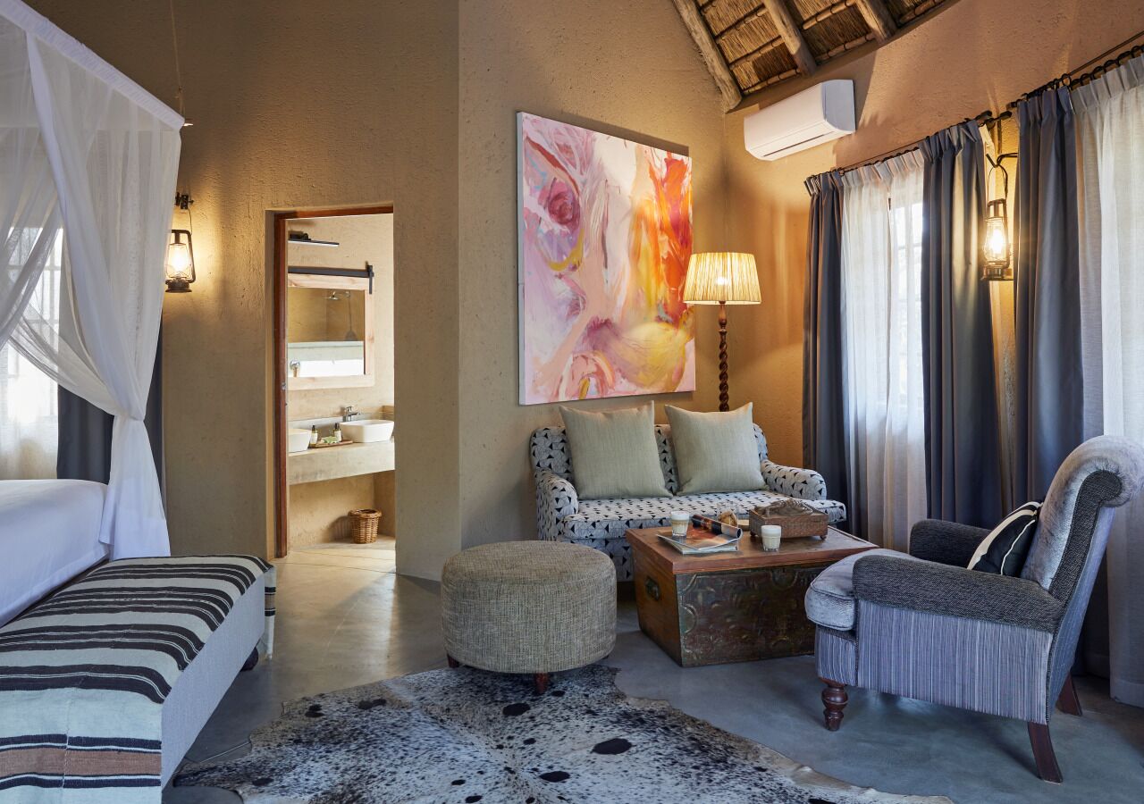 A spacious room with a bed, a sofa, a chair, and a coffee table. There is a painting on the wall and a lamp on the table.