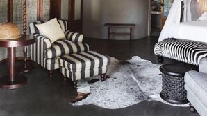 A room with a high ceiling, a bed with a canopy, striped armchairs, a lamp, a wall-mounted antler, and a zebra-patterned rug. - Motswari Private Game Reserve Resort by NEWMARK (Bushbuckridge)