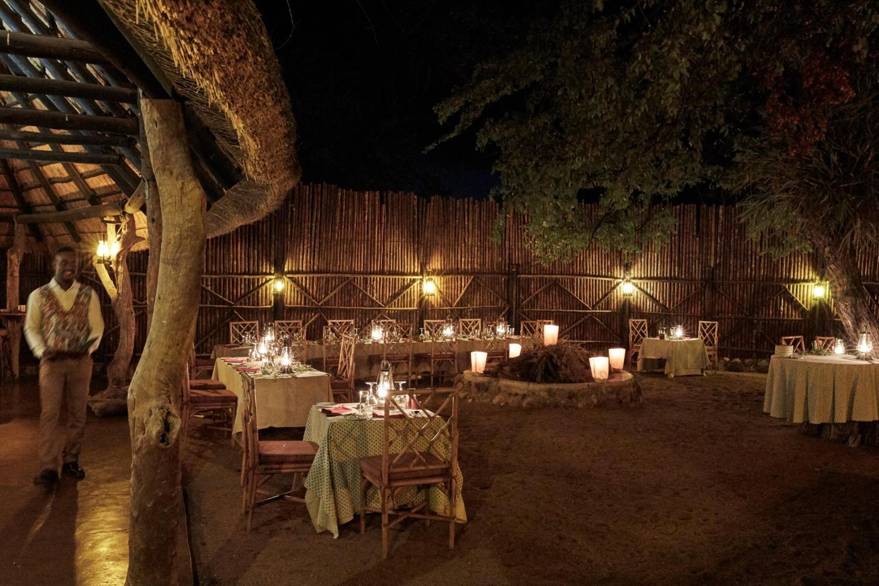 An outdoor dining area with tables set for a meal, surrounded by a rustic wooden fence and illuminated by candles.