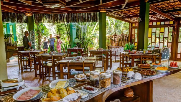 Restaurante com mesas e cadeiras de madeira, buffet variado e vista para um jardim tropical.