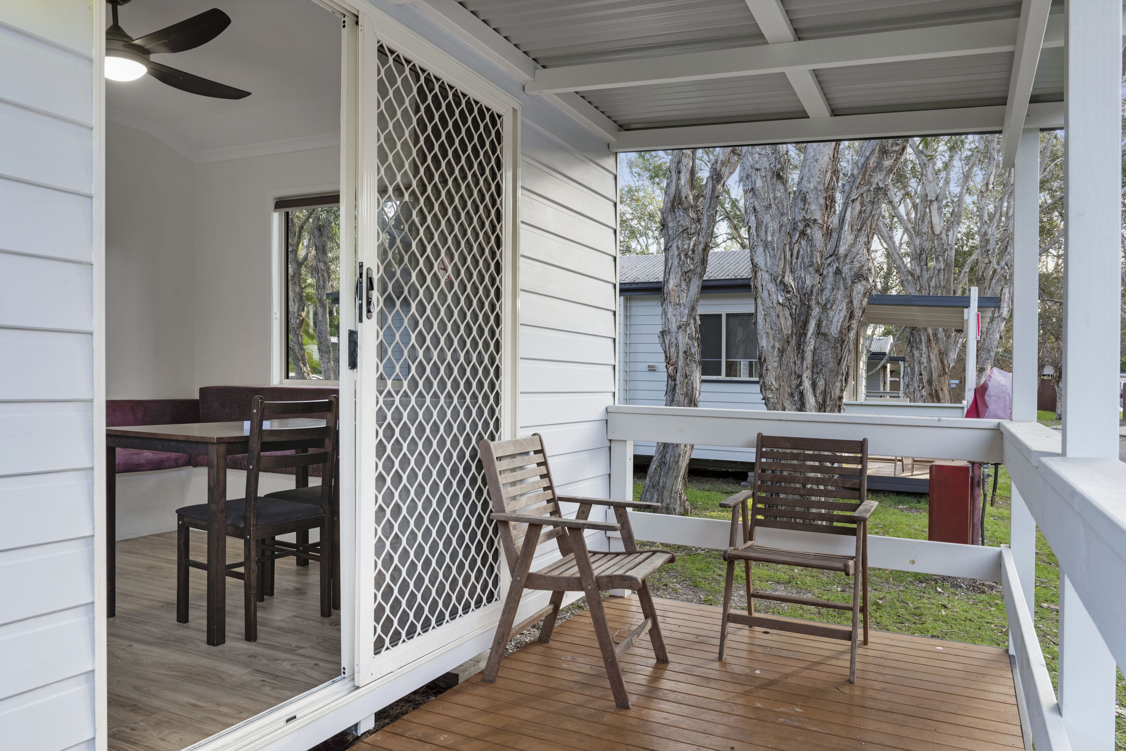 A covered porch with wooden chairs and a table, a screened area, and a view of trees and a building in the background.