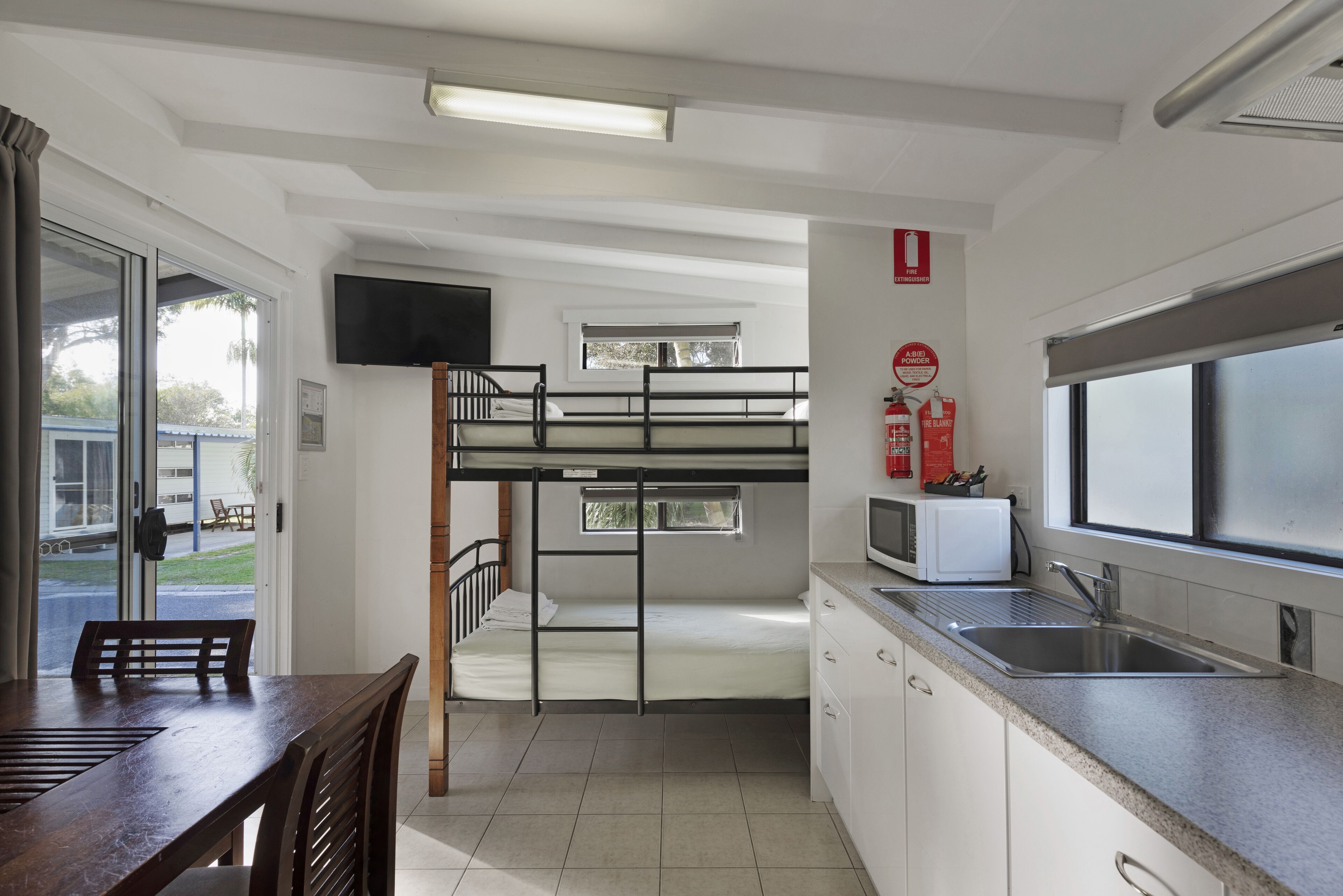 Een slaapkamer met stapelbedden, een keukenhoek met een magnetron, spoelbak en kastjes, en een eettafel met stoelen.